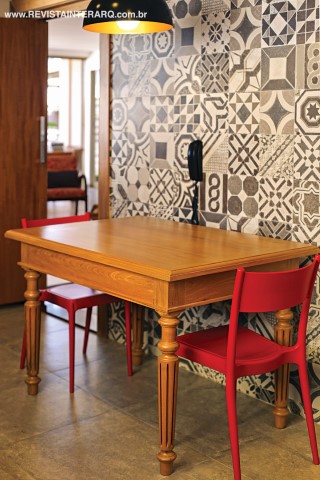 Detalhe da composição da cozinha com parede revestida com porcelanato estilo ladrilho hidráulico, mesa de madeira de demolição (Ismael Gabriel Marcenaria) e cadeiras vermelhas