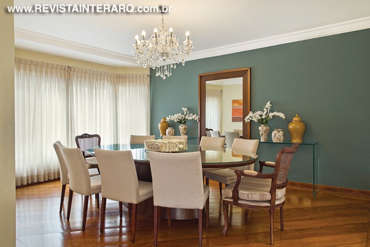 Paredes verdes e cortinas de linho envolvem o jantar em uma atmosfera delicada, acima da mesa oval em ébano e poltronas estofadas, o lustre de cristais é puro luxo