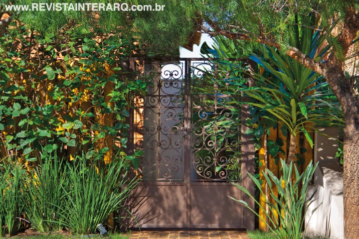 A entrada lateral da casa traz um portão de ferro com ares de antigamente, cercado por Moreias e Strelitzias