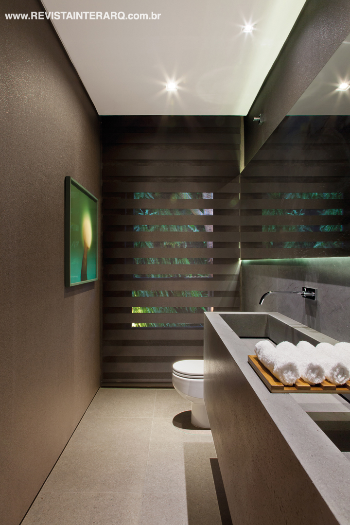 Espelho e bancada de mármore acompanham a extensão da parede do lavabo com papel de parede escuro e luminotécnica intimista. O quadro é de Alberto Oliveira