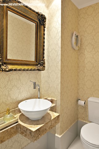 O lavabo traz papel de parede dourado adamascado (ACB Casa), bancada em mármore Crema Marfil e espelho com moldura dourada