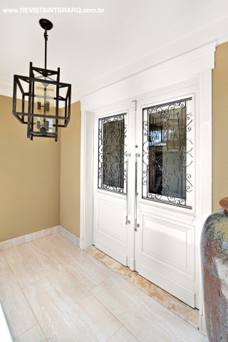 A beleza da porta de entrada em laca branca com gradis arabescados é complementada pelo lustre em ferro da Luminá Iluminação. O piso é em porcelanato Travertino da Portobello Shop
