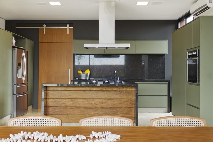 A cozinha mescla planejados na cor verde oliva com granito preto e madeira de demolição. O layout limpo favorece a integração com a varanda gourmet